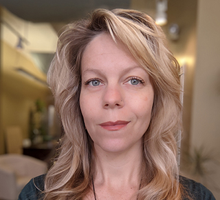 This image is a tight crop photograph of Deanna Lorianna: a white woman with gray eyes, a narrow face, and a tall neck. Her blonde hair is shoulder length. She stands in her office space and is happy with a grin on her face. The background of the image is blurry.