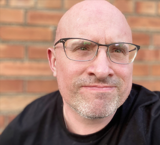 This image is a tight crop photograph of John O'Neill: a white mid-age male with a black shirt and black frame glasses. His head is shaved, and his eyes are pointing upright. John stands in front of a red brick wall.